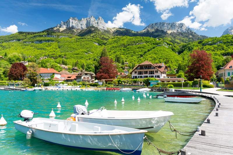 Vue depuis un pont sur le lac d'Annecy, avec des bateaux à quai, des maisons savoyardes et les montagnes en arrière-plan sous un ciel bleu.