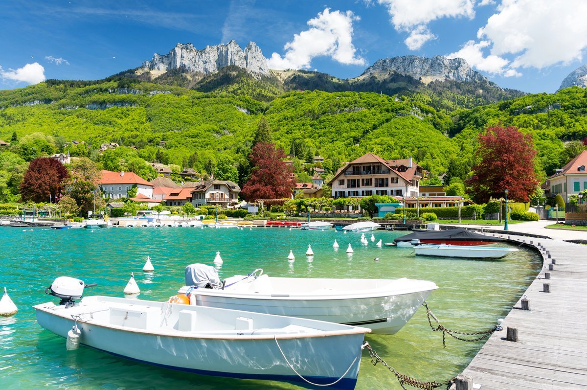 Vue depuis un pont sur le lac d'Annecy, avec des bateaux à quai, des maisons savoyardes et les montagnes en arrière-plan sous un ciel bleu.