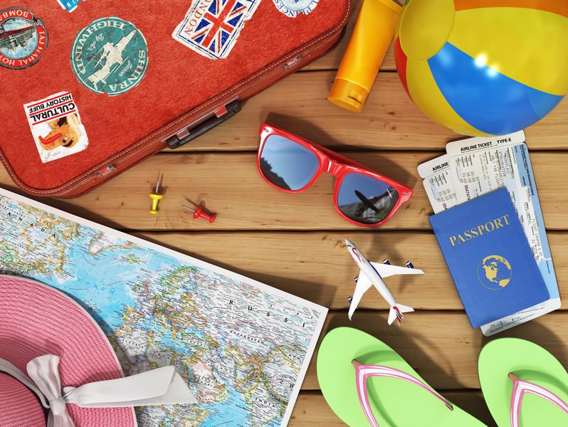 Une valise rouge décorée de plusieurs autocollants de pays, accompagnée d’un passeport, d’un billet d’avion, d’une carte du monde, de lunettes de soleil rouges et d’autres articles de voyage disposés sur une table en bois.