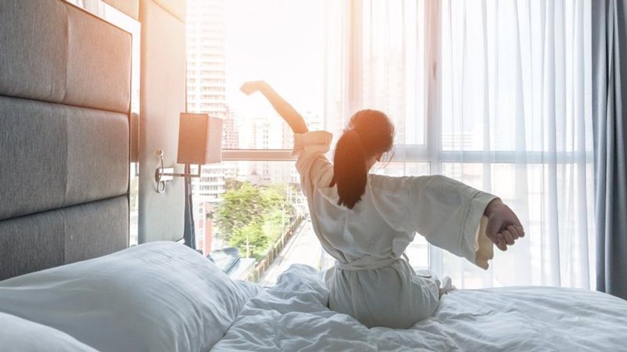 Femme en peignoir s’étirant au réveil dans une chambre d’hôtel lumineuse avec vue sur la ville.