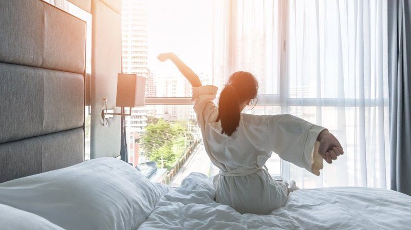 Femme en peignoir s’étirant au réveil dans une chambre d’hôtel lumineuse avec vue sur la ville.