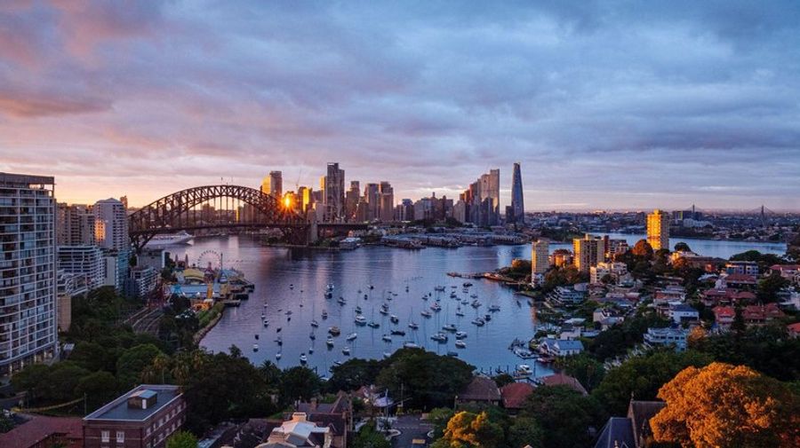 Vue panoramique sur le Harbour Bridge et le centre-ville de Sydney au coucher du soleil, avec des voiliers dans la baie.