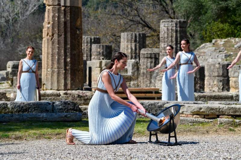 Cérémonie traditionnelle avec des prêtresses allumant la flamme olympique sur le site historique d’Olympie, entourées de ruines antiques.