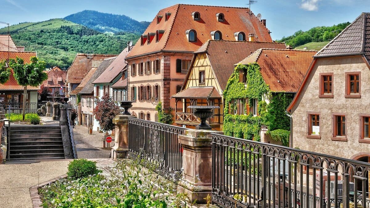 Maisons alsaciennes colorées et façades à colombages dans un village fleuri entouré de collines verdoyantes.
