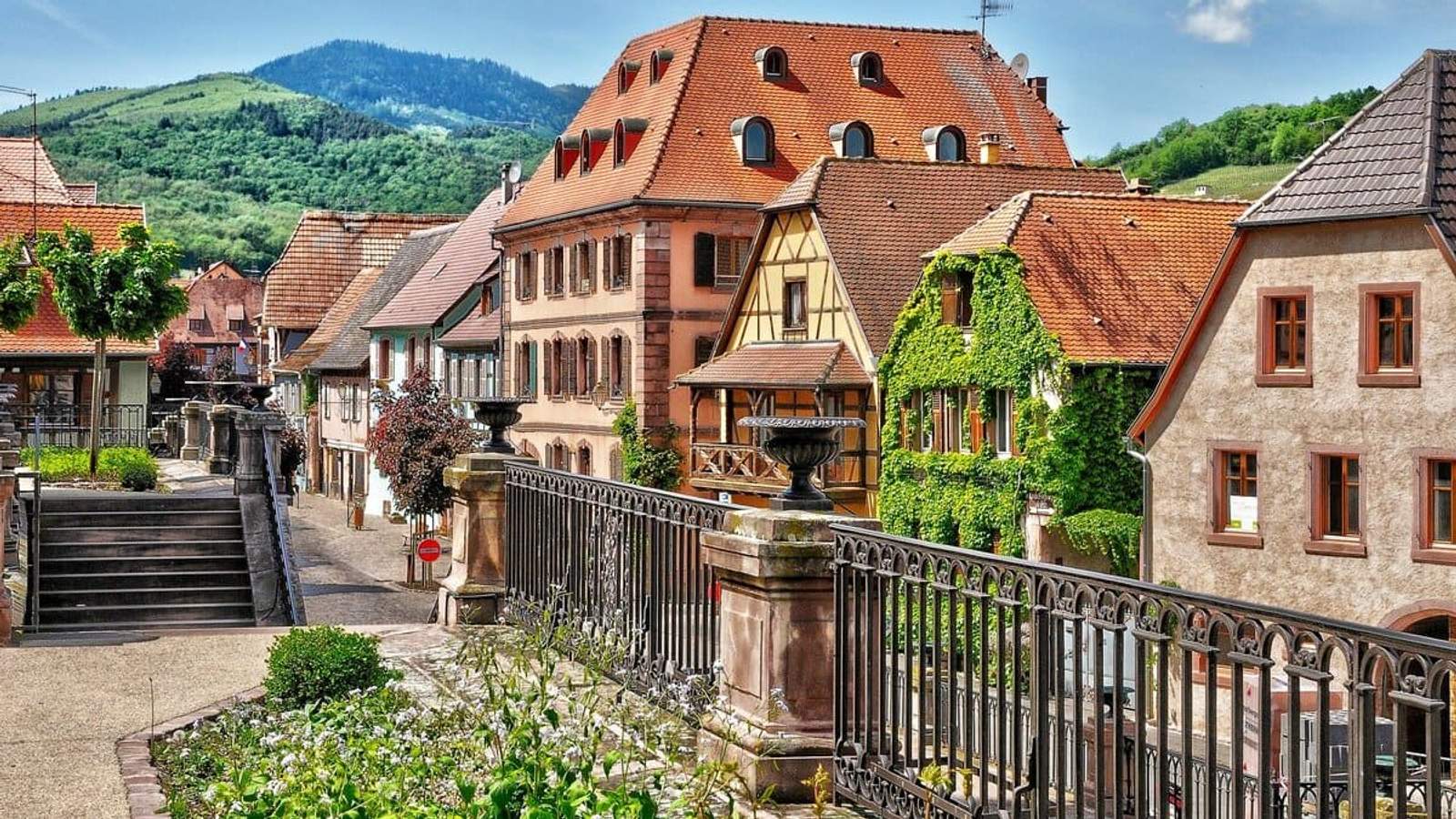 Maisons alsaciennes colorées et façades à colombages dans un village fleuri entouré de collines verdoyantes.