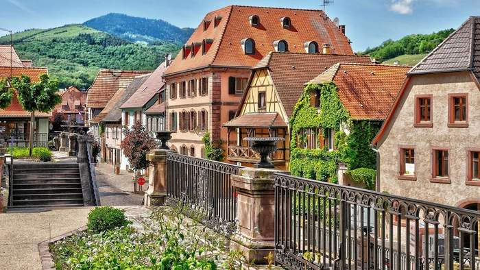 Maisons alsaciennes colorées et façades à colombages dans un village fleuri entouré de collines verdoyantes.