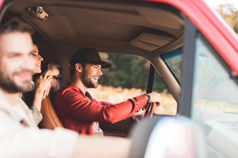 Un groupe d'amis souriants à bord d'un véhicule, profitant d'un road trip à travers la campagne. Le conducteur, portant une casquette, est concentré et détendu, tandis que les passagers partagent un moment de convivialité, reflétant l'esprit de voyage et d'aventure.