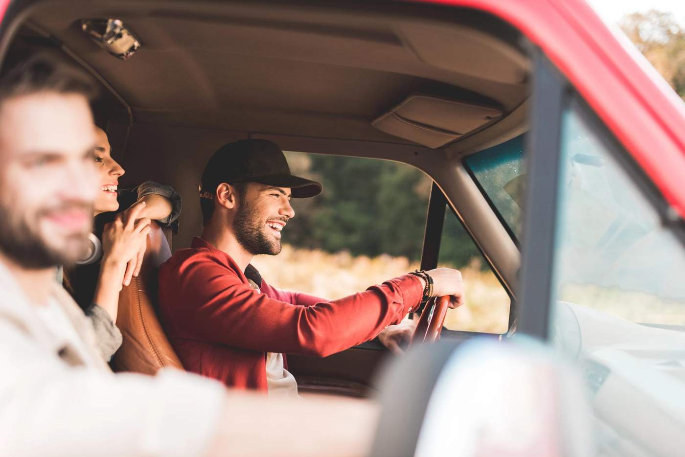 Un groupe d'amis souriants à bord d'un véhicule, profitant d'un road trip à travers la campagne. Le conducteur, portant une casquette, est concentré et détendu, tandis que les passagers partagent un moment de convivialité, reflétant l'esprit de voyage et d'aventure.
