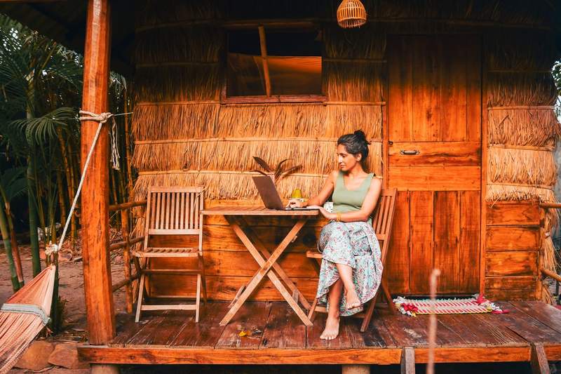 Community manager digital nomade travaillant sur son ordinateur portable, assise sur la terrasse en bois d'une cabane tropicale au toit de chaume