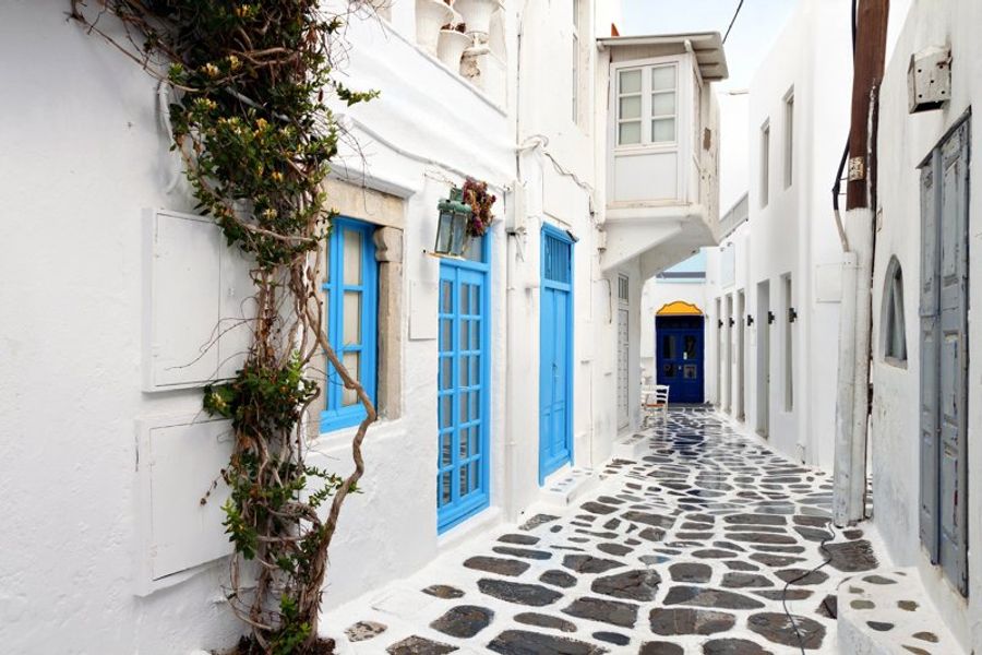 Ruelle typique de Mykonos avec des murs blancs, des portes et fenêtres bleues, et un sol pavé.