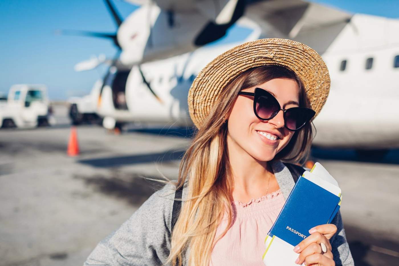 Jeune voyageuse souriante avec un passeport et un billet d’avion à la main devant un avion sur le tarmac.