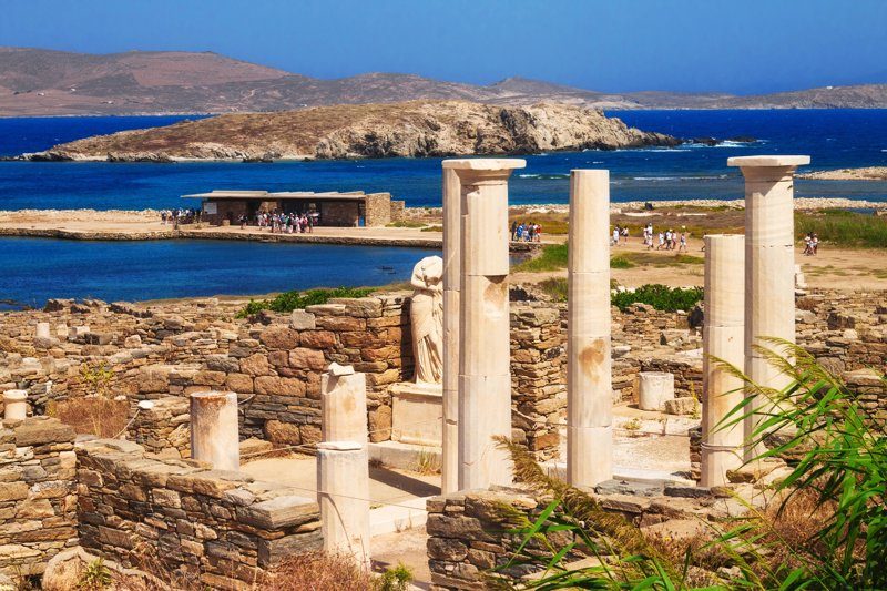 Ruines antiques de l’île de Délos avec des colonnes blanches et une vue sur la mer Égée.
