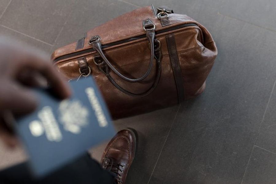 Sac de voyage en cuir marron posé au sol avec un passeport tenu à la main