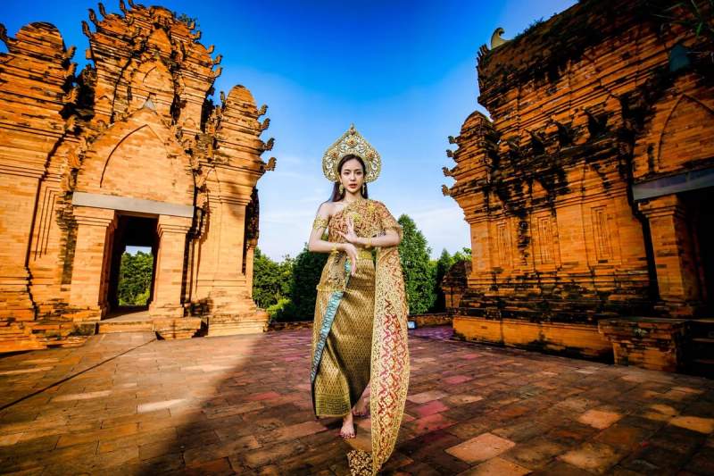 Une femme en tenue traditionnelle thaïlandaise posant devant des temples anciens en briques rouges, sous un ciel bleu éclatant.