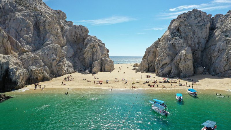 Vue de la plage de Cabo San Lucas, entourée de formations rocheuses impressionnantes avec des visiteurs et des bateaux.