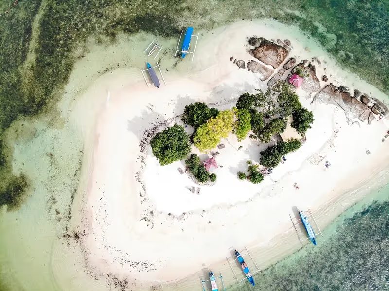 Une vue aérienne d'une l'ile Meno avec des bateaux à voile bleus amarrés près de plages de sable blanc, entourée par des eaux peu profondes.