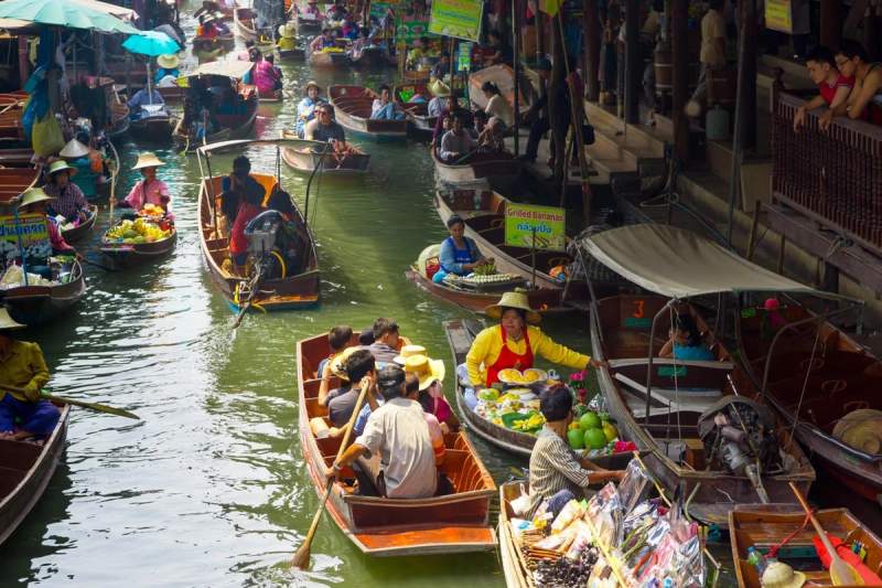 Scène animée d’un marché flottant en Thaïlande, avec des bateaux remplis de fruits, plats locaux et produits artisanaux, entourés de marchands et de visiteurs.