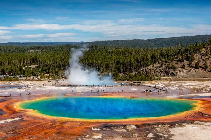 Vue aérienne spectaculaire de la Grand Prismatic Spring dans le parc national de Yellowstone, avec ses couleurs vives entourées de visiteurs et d'une forêt dense en arrière-plan.