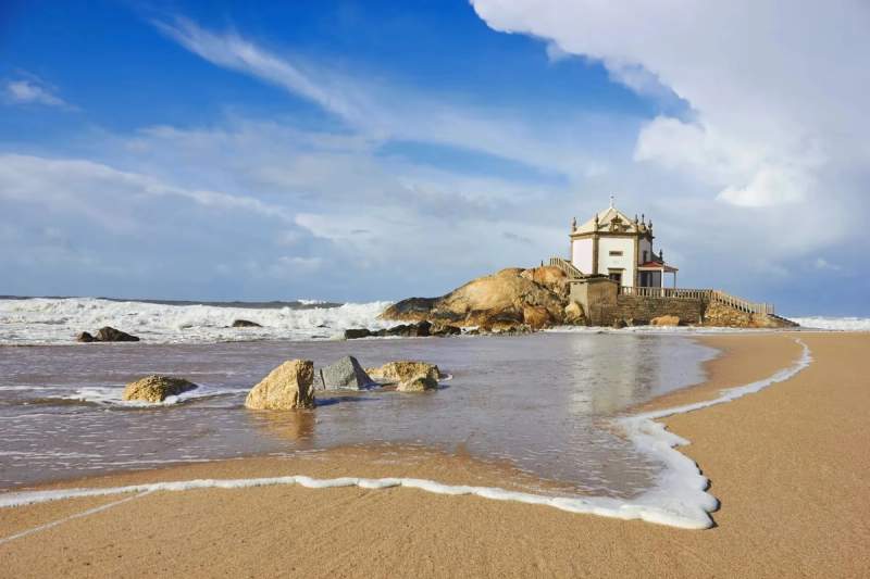 Vue de la chapelle du Senhor da Pedra sur la plage, avec des vagues se brisant sur les rochers sous un ciel bleu éclatant.