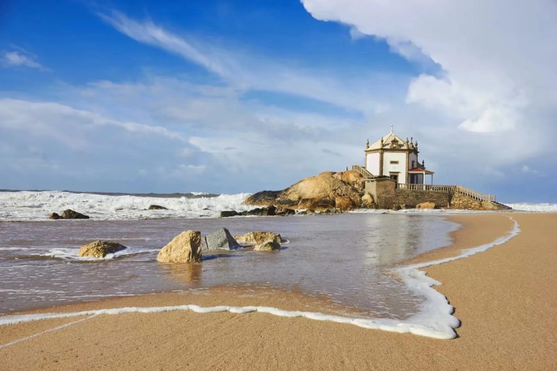 Vue de la chapelle du Senhor da Pedra sur la plage, avec des vagues se brisant sur les rochers sous un ciel bleu éclatant.