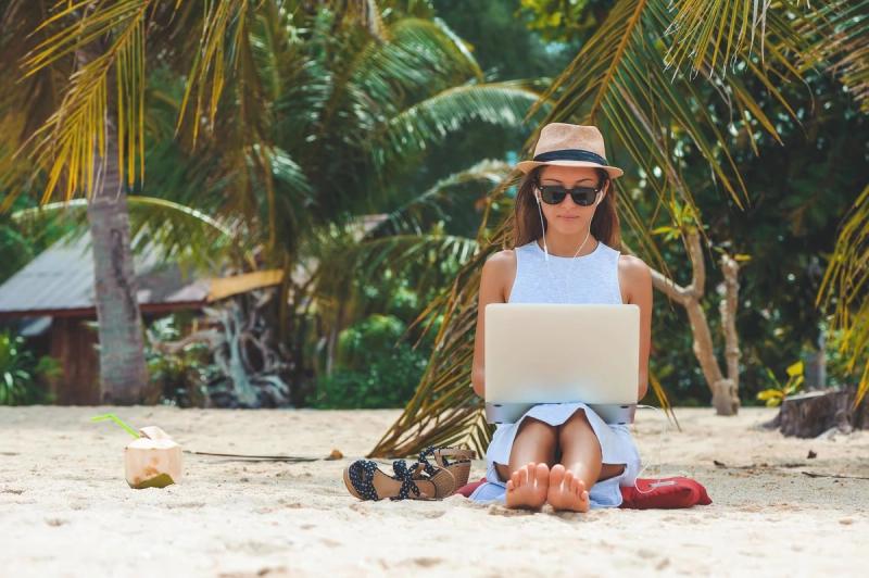 Femme freelance travaillant sur un ordinateur portable assise sur une plage tropicale, entourée de palmiers, avec une noix de coco à proximité. Scène typique d'un nomade digital profitant du travail à distance en pleine nature.