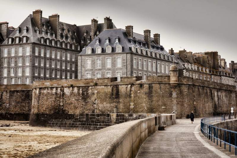 Remparts de la ville close de Saint-Malo en Bretagne, avec ses bâtiments en granit et une promenade piétonne longeant la mer sous un ciel gris.