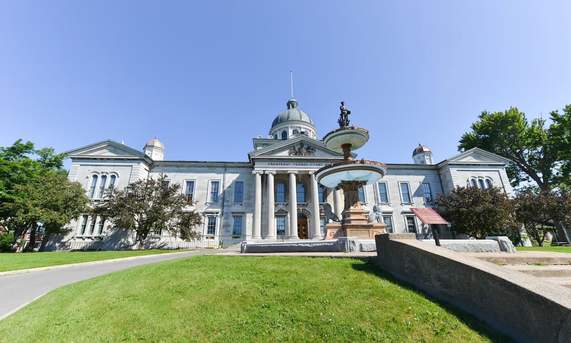 Façade du Frontenac County Court à Kingston avec sa fontaine historique et son architecture en pierre calcaire sous un ciel bleu.