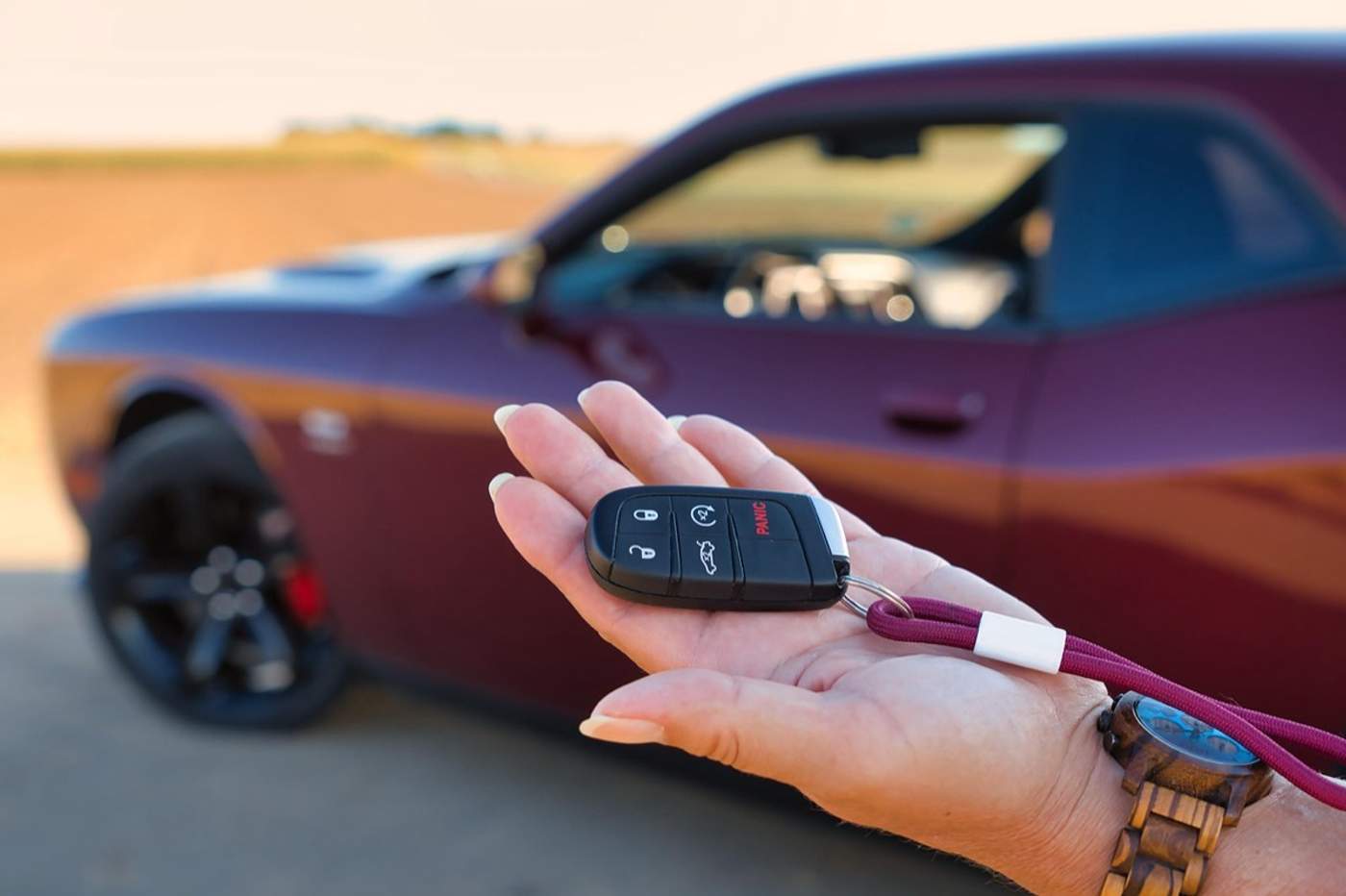 Main tendue présentant la clé d'une voiture de location, devant un coupé bordeaux garé au bord d'un champ.
