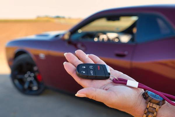 Main tendue présentant la clé d'une voiture de location, devant un coupé bordeaux garé au bord d'un champ.