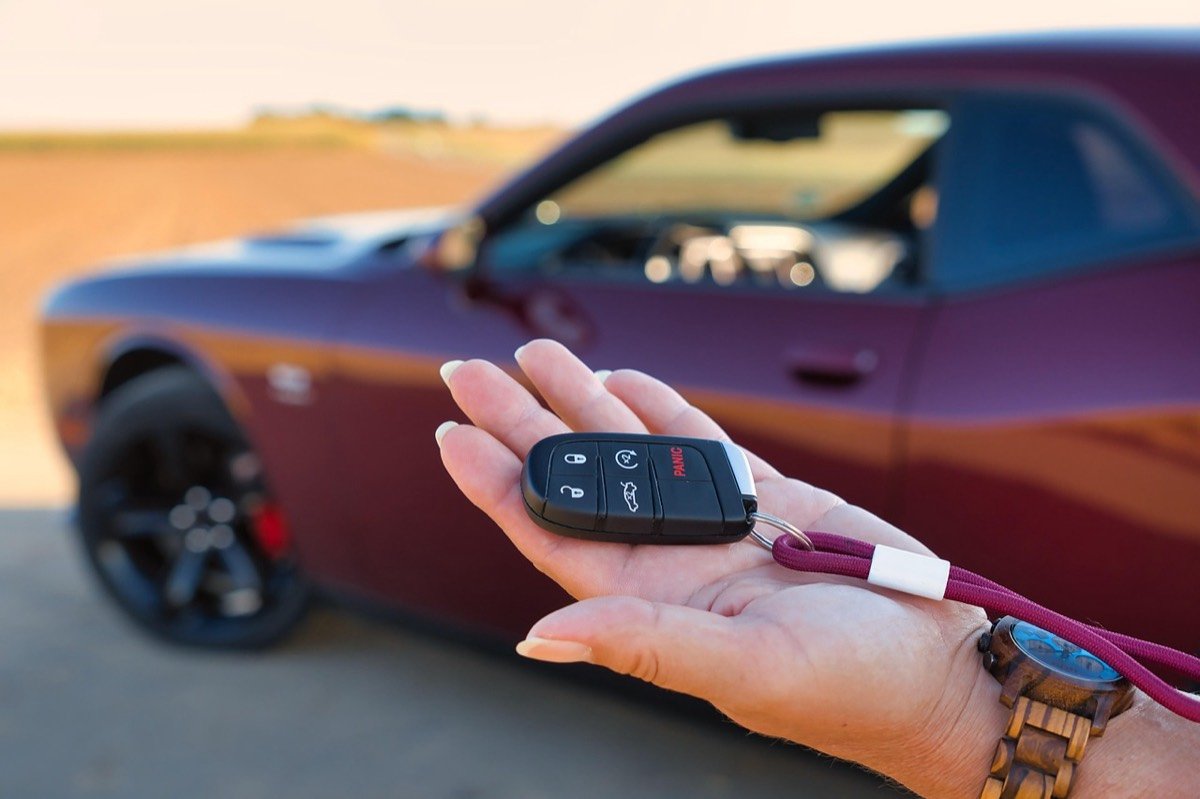 Main tendue présentant la clé d'une voiture de location, devant un coupé bordeaux garé au bord d'un champ.
