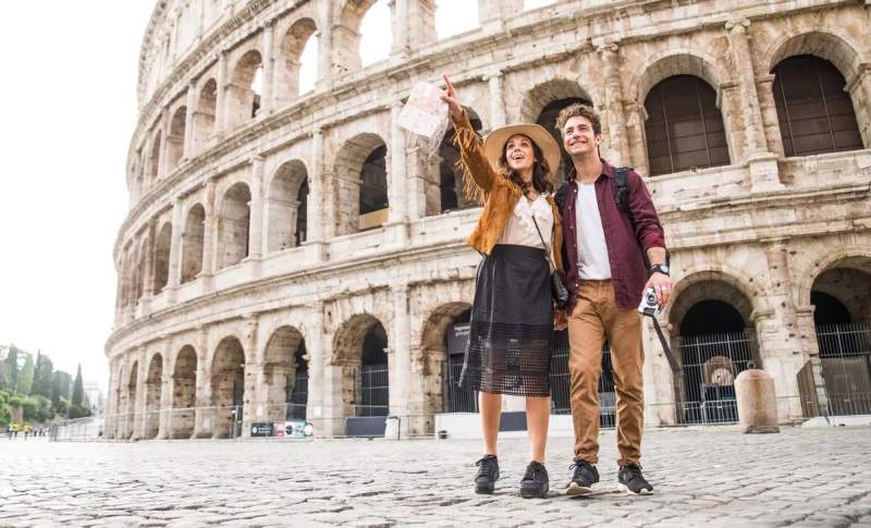 Couple de voyageurs souriants visitant le Colisée à Rome, tenant une carte et un appareil photo.