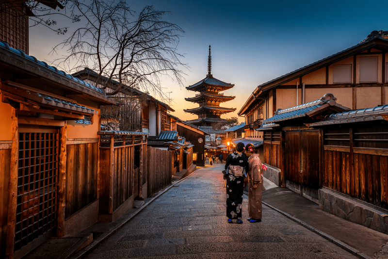 Rue traditionnelle de Kyoto au coucher du soleil avec deux femmes en kimono et une pagode en arrière-plan.