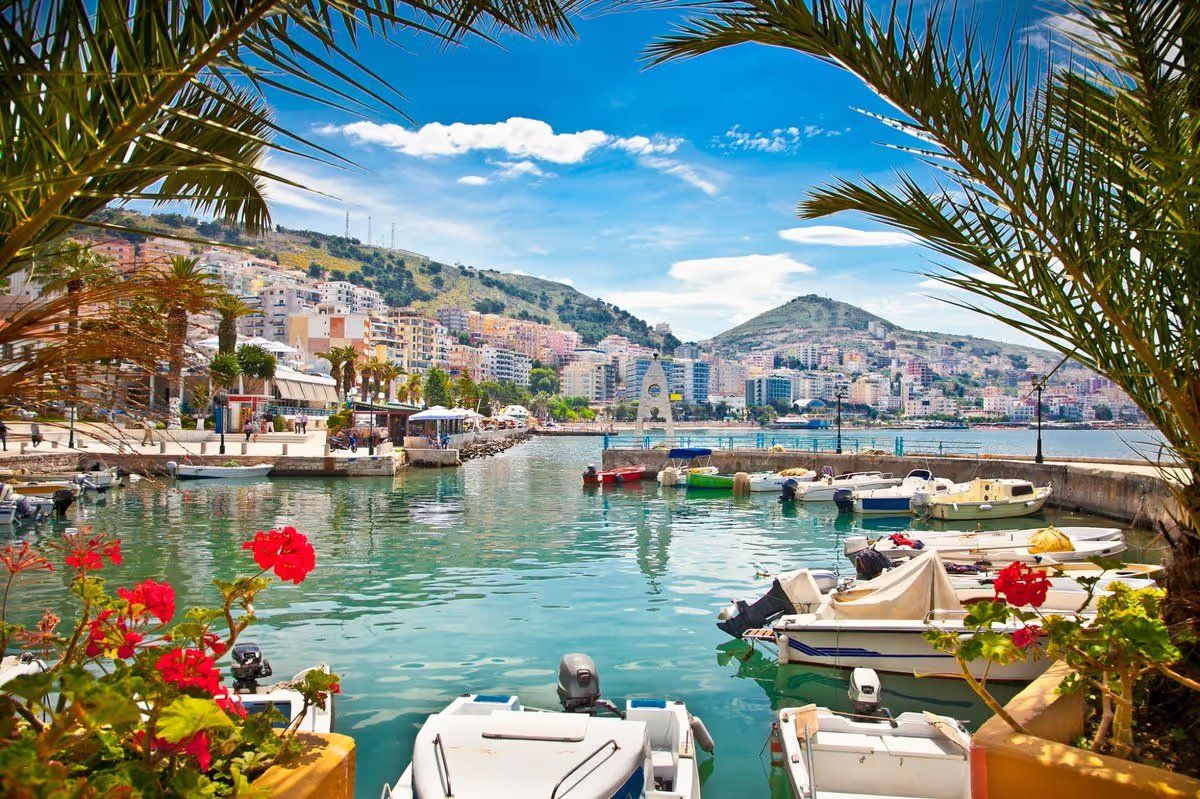 Port de Saranda en Albanie avec bateaux de pêche, palmiers et immeubles colorés en bord de mer sous un ciel bleu