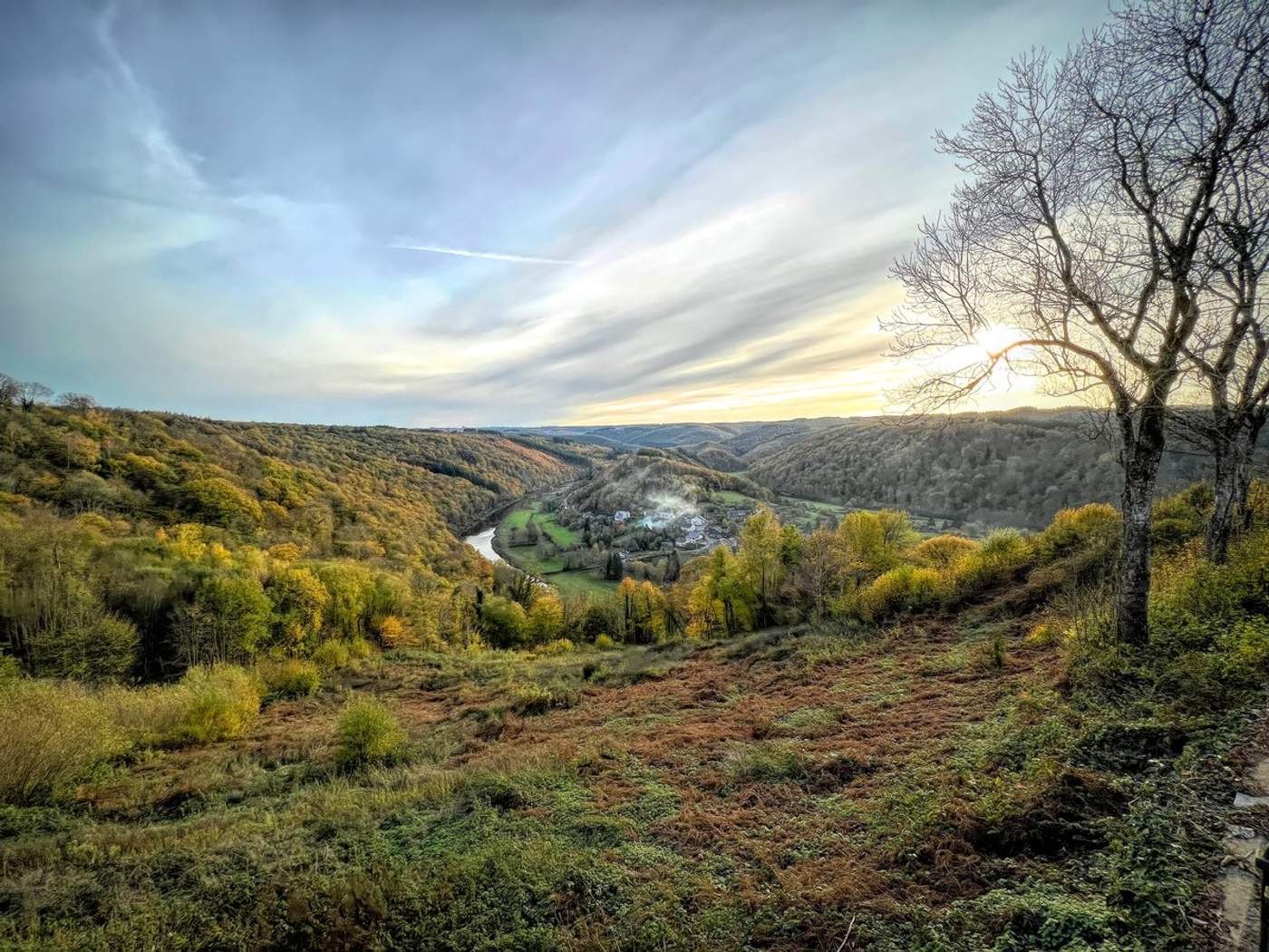 Vue panoramique sur une vallée boisée en automne avec une rivière sinueuse et un village au coucher du soleil dans les Ardennes