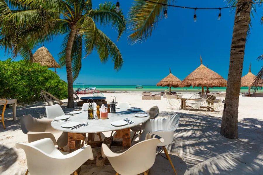 Une table dressée pour un déjeuner est installée sous des palmiers sur une plage de sable blanc, avec une vue dégagée sur la mer turquoise à Holbox.