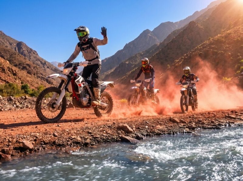 Trois pilotes à moto enduro traversant une piste près d’un oued dans l’Atlas marocain