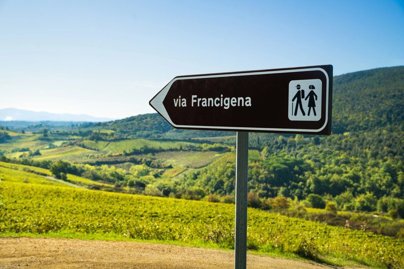 Panneau indiquant la Via Francigena, avec un paysage toscan verdoyant en arrière-plan.
