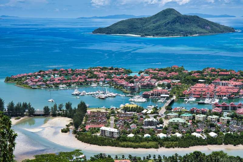 Vue aérienne d'une marina luxueuse aux Seychelles avec de nombreux bateaux amarrés, entourée de villas aux toits rouges et de bâtiments résidentiels, avec une île verdoyante et l'océan bleu en arrière-plan.