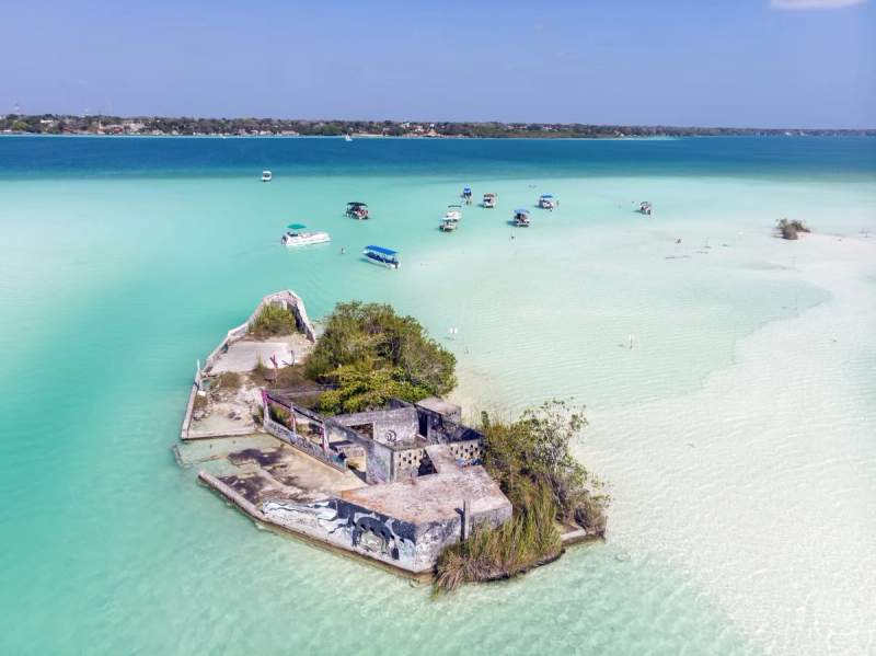 Vue aérienne d'une petite île abandonnée dans la lagune de Bacalar, entourée de bateaux dans une eau turquoise claire.