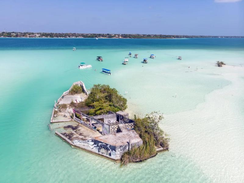 Vue aérienne d'une petite île abandonnée dans la lagune de Bacalar, entourée de bateaux dans une eau turquoise claire.