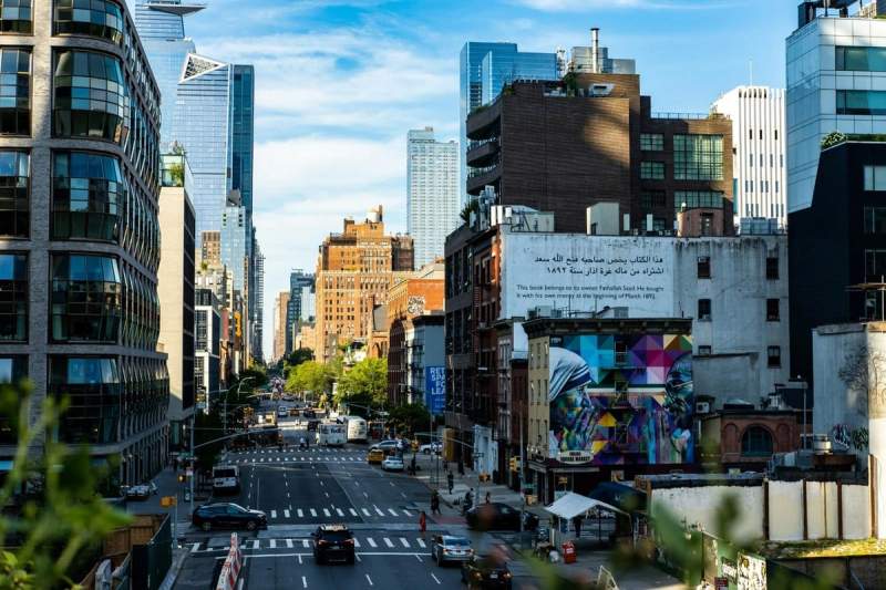 Vue animée d’une avenue à Manhattan bordée de gratte-ciel modernes et d’immeubles en briques, avec une fresque colorée au premier plan.
