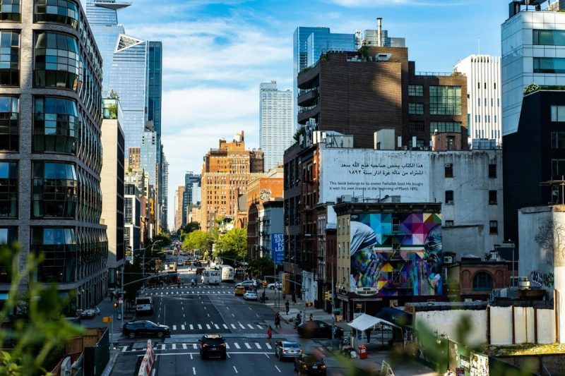 Vue animée d’une avenue à Manhattan bordée de gratte-ciel modernes et d’immeubles en briques, avec une fresque colorée au premier plan.