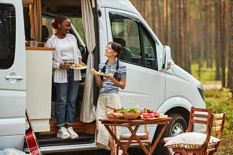 Deux femmes partagent un repas devant un van aménagé lors d’un road trip en pleine forêt