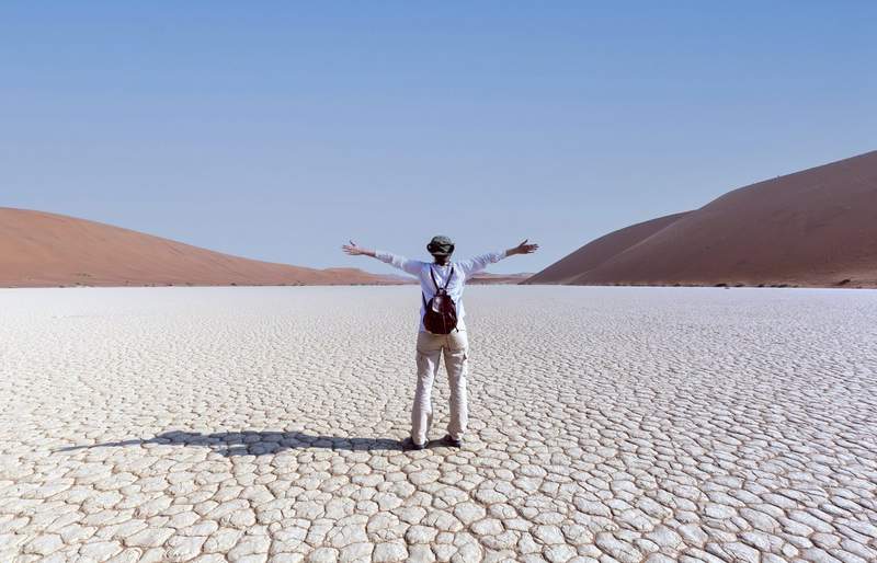 Voyageur de dos, bras ouverts, admirant l'immensité du désert de Namibie avec ses dunes rouges et son sol craquelé sous un ciel bleu dégagé.