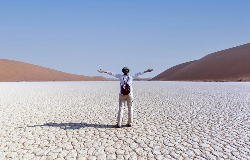 Voyageur de dos, bras ouverts, admirant l'immensité du désert de Namibie avec ses dunes rouges et son sol craquelé sous un ciel bleu dégagé.