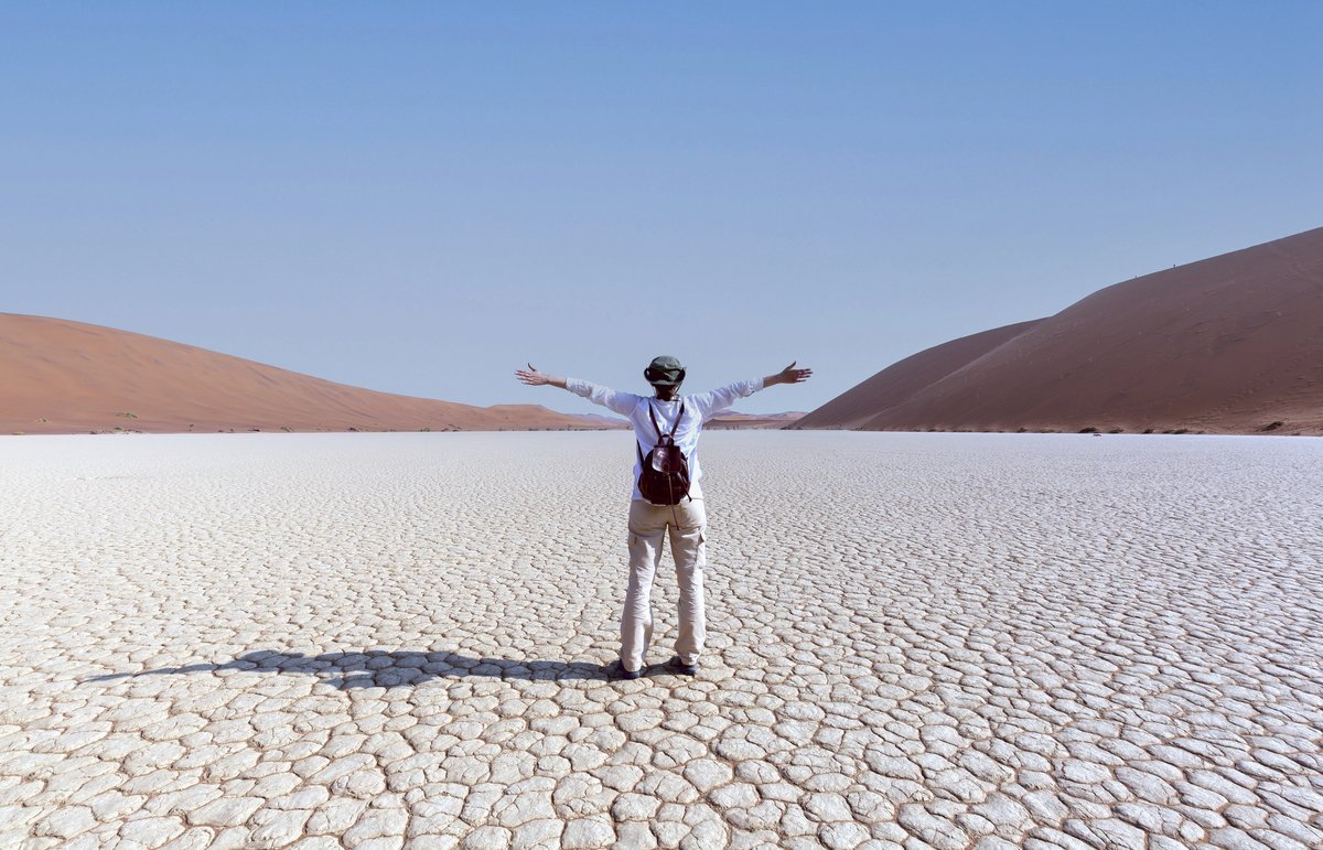 Voyageur de dos, bras ouverts, admirant l'immensité du désert de Namibie avec ses dunes rouges et son sol craquelé sous un ciel bleu dégagé.