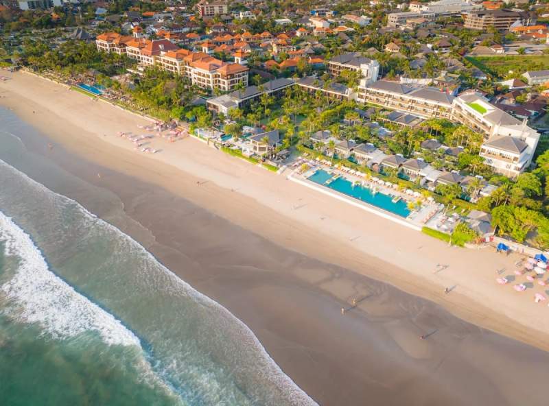 Vue aérienne d'une plage à Seminyak, Bali, avec des complexes hôteliers en bord de mer et des piscines surplombant l'océan.