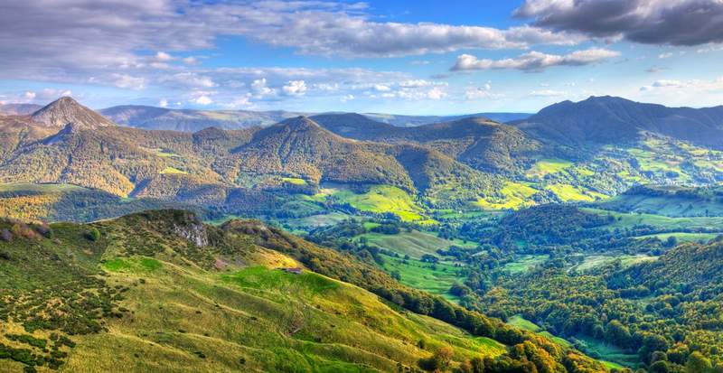 Vue panoramique sur les reliefs verdoyants et volcaniques du Massif Central en Auvergne, sous un ciel partiellement nuageux.