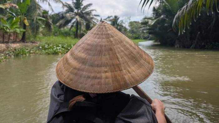 Vue arrière de Maud portant un chapeau conique sur une barque naviguant dans un canal bordé de palmiers à Ben Tre au Vietnam