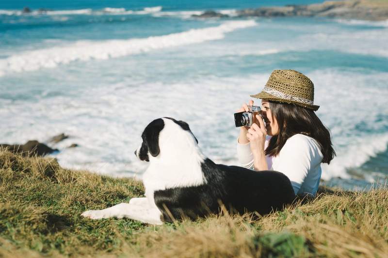 Voyageuse prenant une photo au bord de la mer avec son chien allongé dans l’herbe, lors d’un séjour en pleine nature.